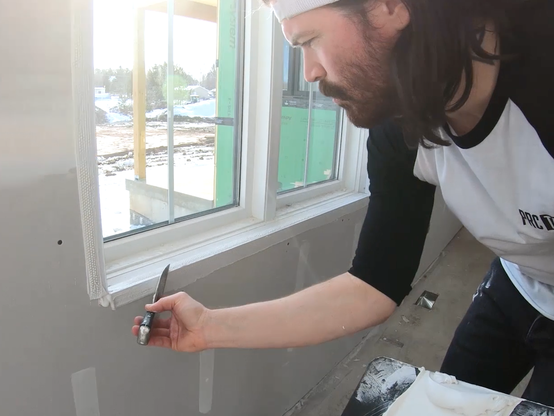 A person applies joint compound on a drywall corner bead next to a window.